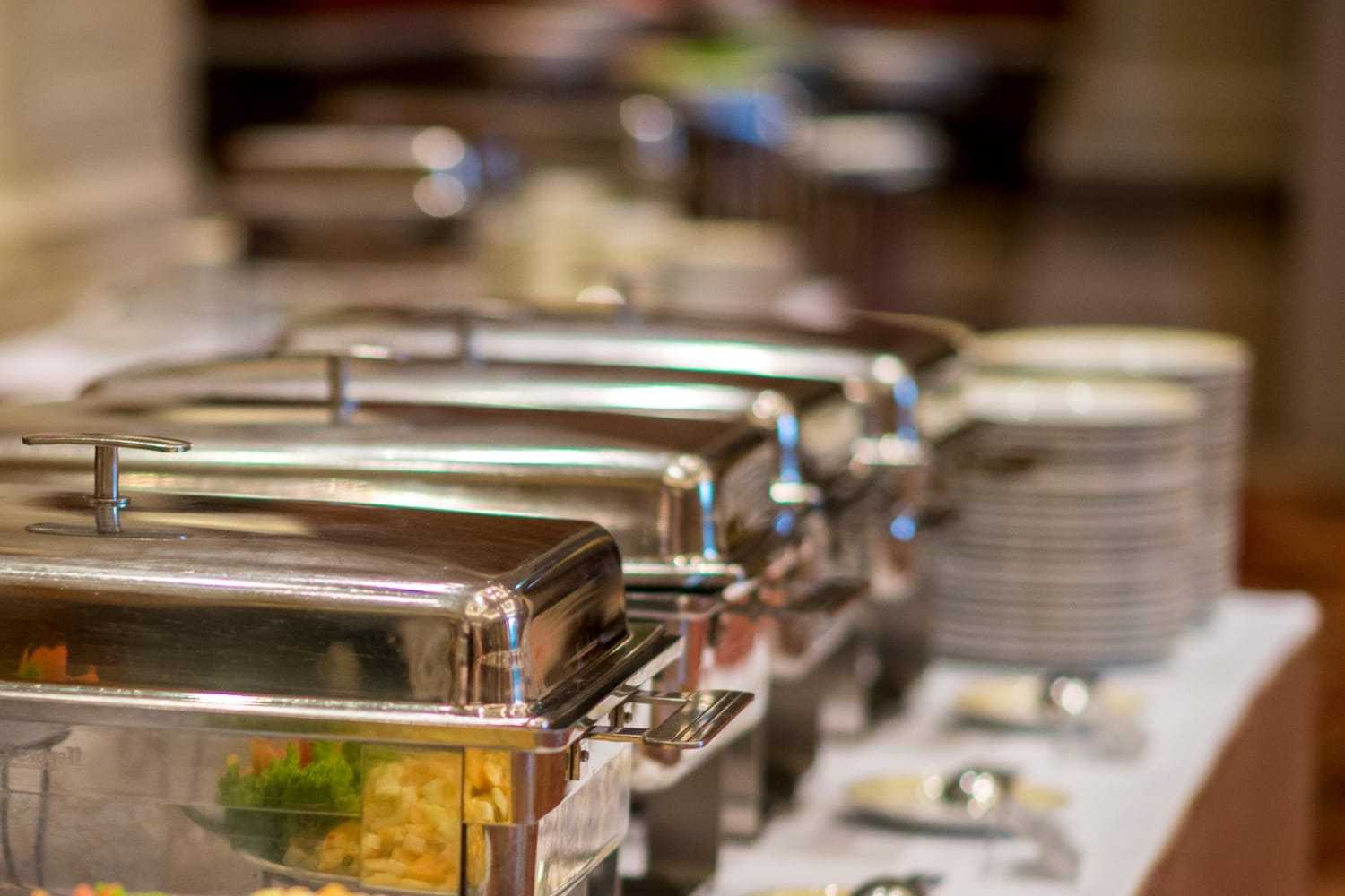 Buffet setup with covered food trays and stacks of plates on a table.