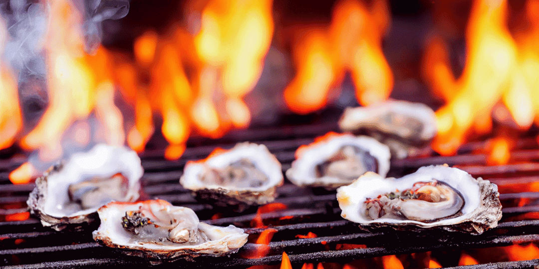 Grilled oysters sizzling on a barbecue with flames in the background.