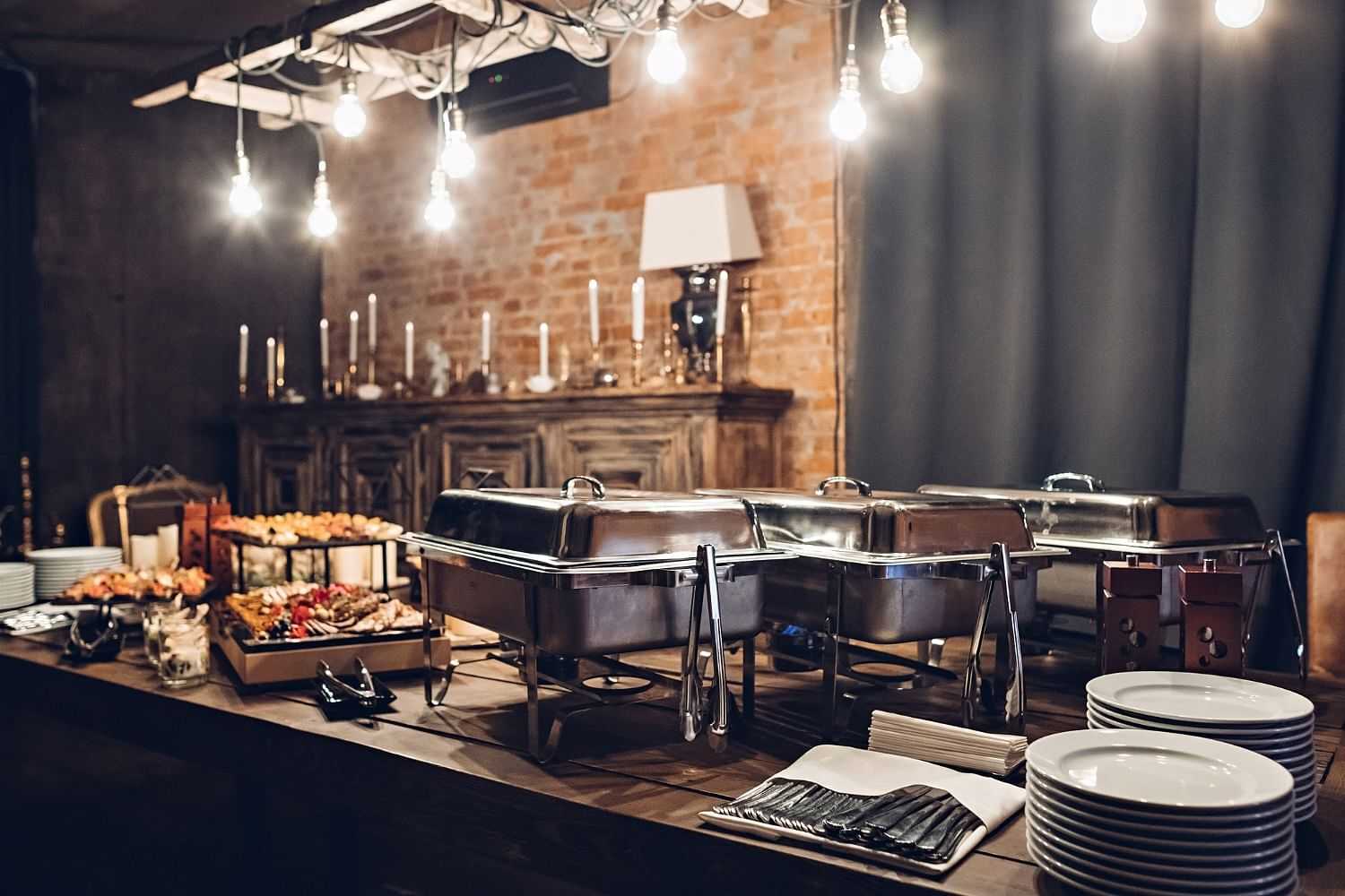 Elegant buffet setup with chafing dishes, assorted food, plates, and dim lighting for an event.