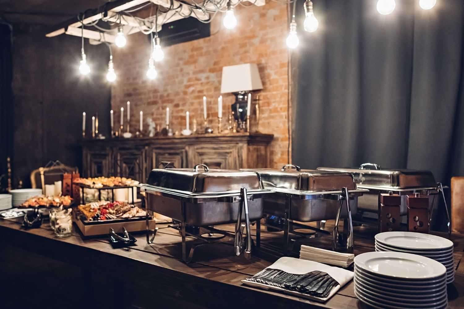Elegant buffet setup with chafing dishes, assorted food, plates, and dim lighting for an event.